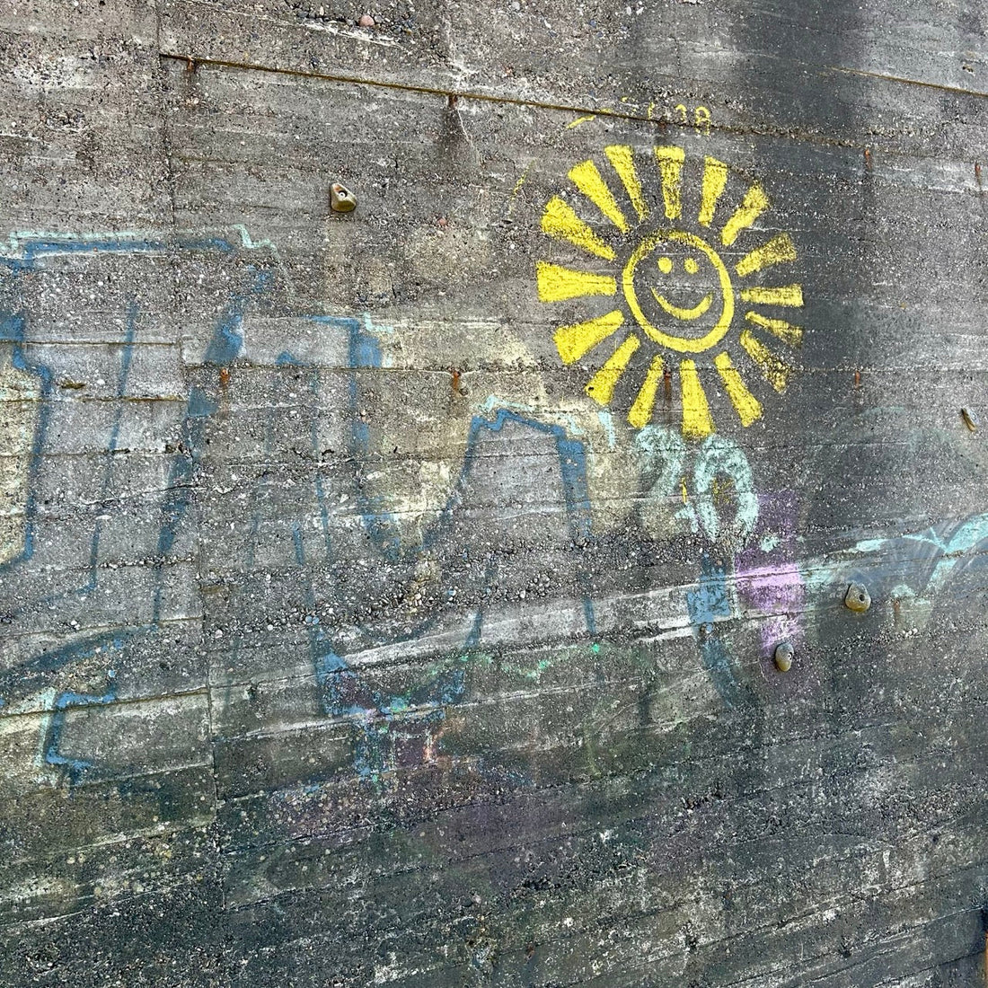 Gelbe Sonne mit lachendem Gesicht als Graffiti auf rauer Betonwand, umgeben von verblassten, bunten Farbspuren