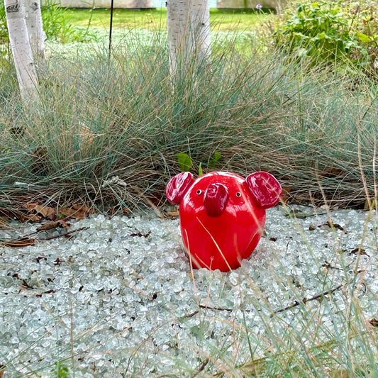 Eine rote Keramikfigur mit großen Ohren und Rüssel steht auf glitzernden Glasscherben, umgeben von hohem Gras und Birken.
