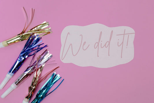 Das Bild zeigt bunte Partytröten auf pinkem Hintergrund mit einem ausgeschnittenen Schild, auf dem „We did it!“ steht – eine festliche und fröhliche Stimmung.