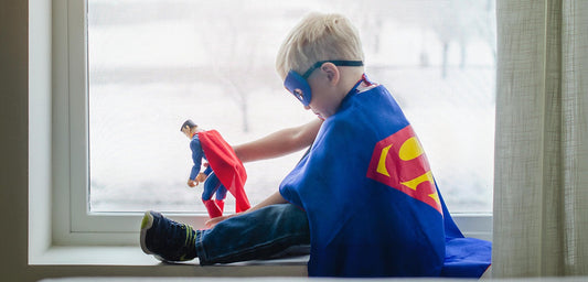 Ein blondes Kind sitzt im Fenster, trägt ein Superman-Cape und eine blaue Maske. Es spielt mit einer Superman-Actionfigur und wirkt konzentriert und fantasievoll.