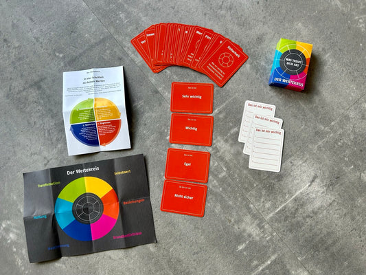 Das Wertekreis-Kartenset liegt ausgebreitet auf grauem Boden: rote Werte- und Bewertungskarten, Anleitungen und eine bunte Box mit der Frage „Was treibt dich an?“.