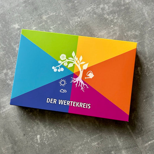 Bunte Spielbox „Der Wertekreis“ mit stilisiertem Baum, Sonne, Wolken und Gießkanne auf grauem Untergrund – symbolisiert Entwicklung und Wertevielfalt.
