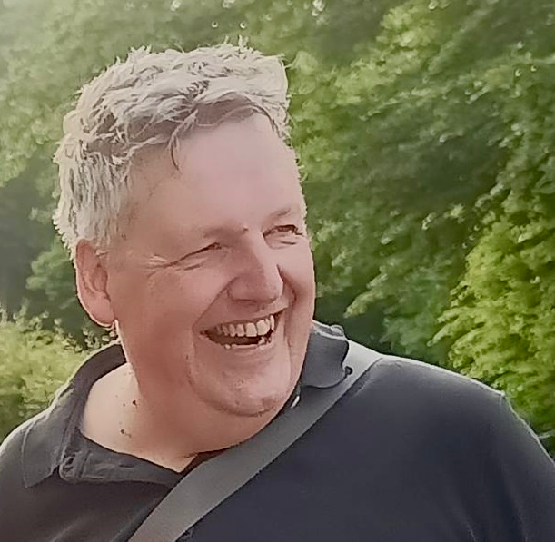 Ein lachender Mann mit grauem Haar und dunklem Polohemd steht im Grünen. Er trägt eine Umhängetasche und wirkt entspannt und lebensfroh.