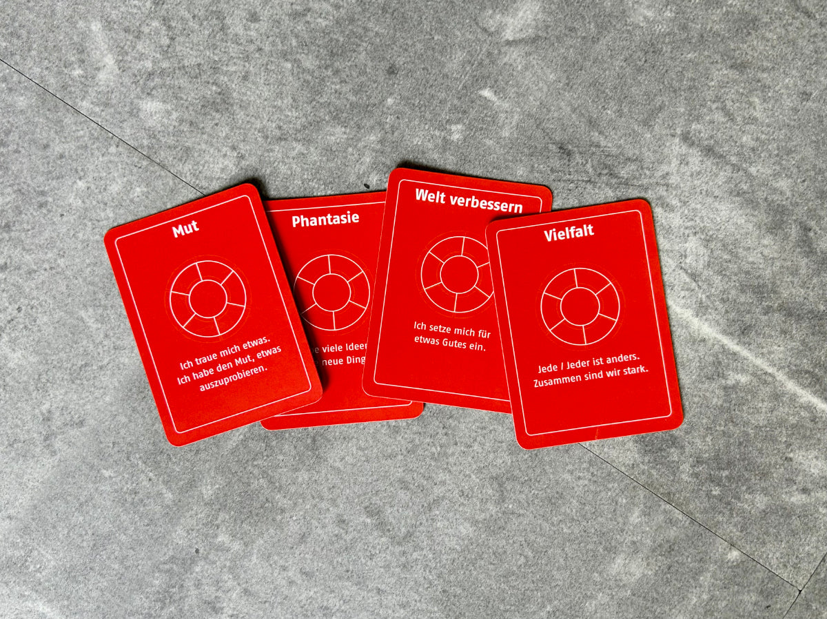 Vier orangefarbene Spielkarten mit den Begriffen Mut, Phantasie, Welt verbessern und Vielfalt liegen leicht überlappend auf einem grau gemusterteten Hintergrund.