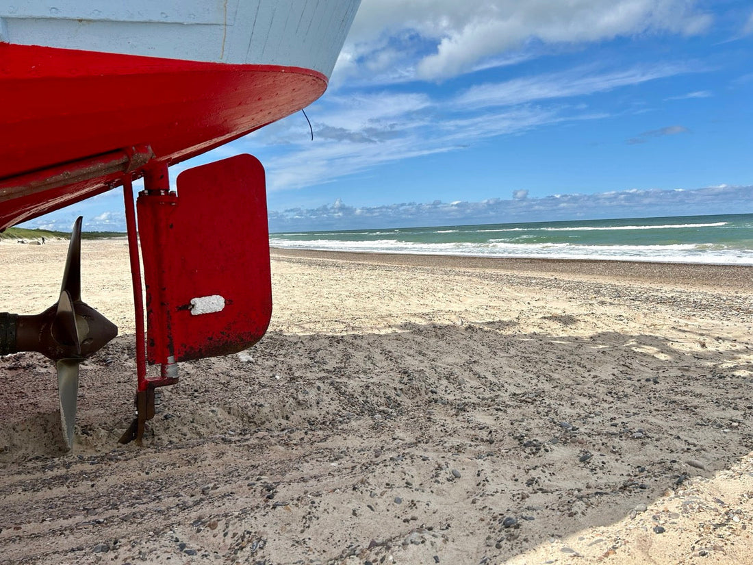 Das Heck eines Fischkutters auf dem Sandstrand mit rotem Ruder und Schiffsschraube. Dahinter das weite Meer, Wellen und ein blauer Himmel mit weißen Wolken.