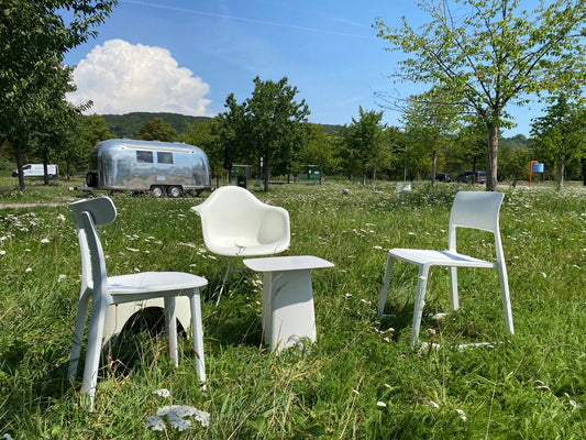 Drei weiße Stühle stehen in einer blühenden Wiese im Kreis um einen kleinen Tisch. Im Hintergrund: Bäume, blauer Himmel und ein silberner Wohnwagen.