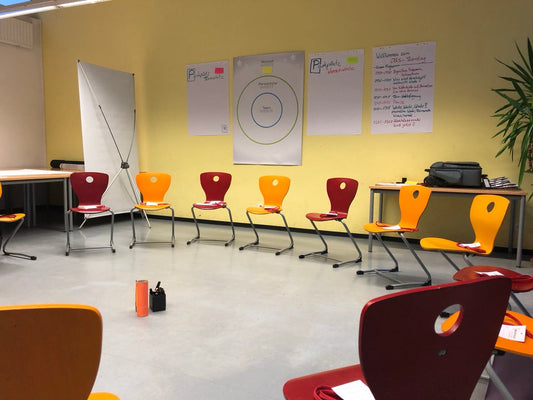 Blick in ein Klassenzimmer. Ein Stuhlkreis mit gelben und roten Holzstühlen, in der Mitte einige Stifte. An der Wand hängen Flipcharts.