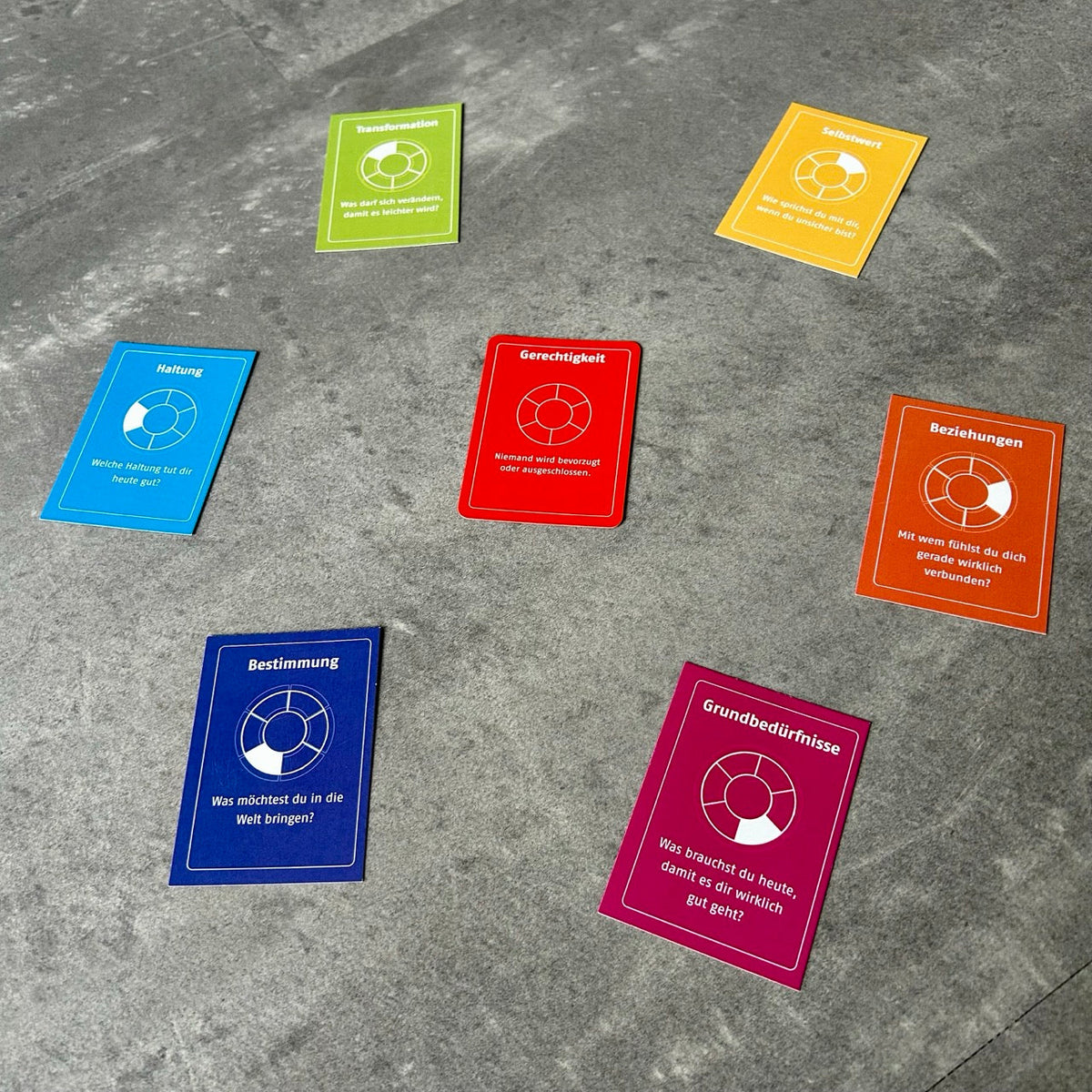 Bunte Wertekarten liegen kreisförmig auf grauem Boden angeordnet. Jede Karte zeigt einen Wert und eine passende Reflexionsfrage.