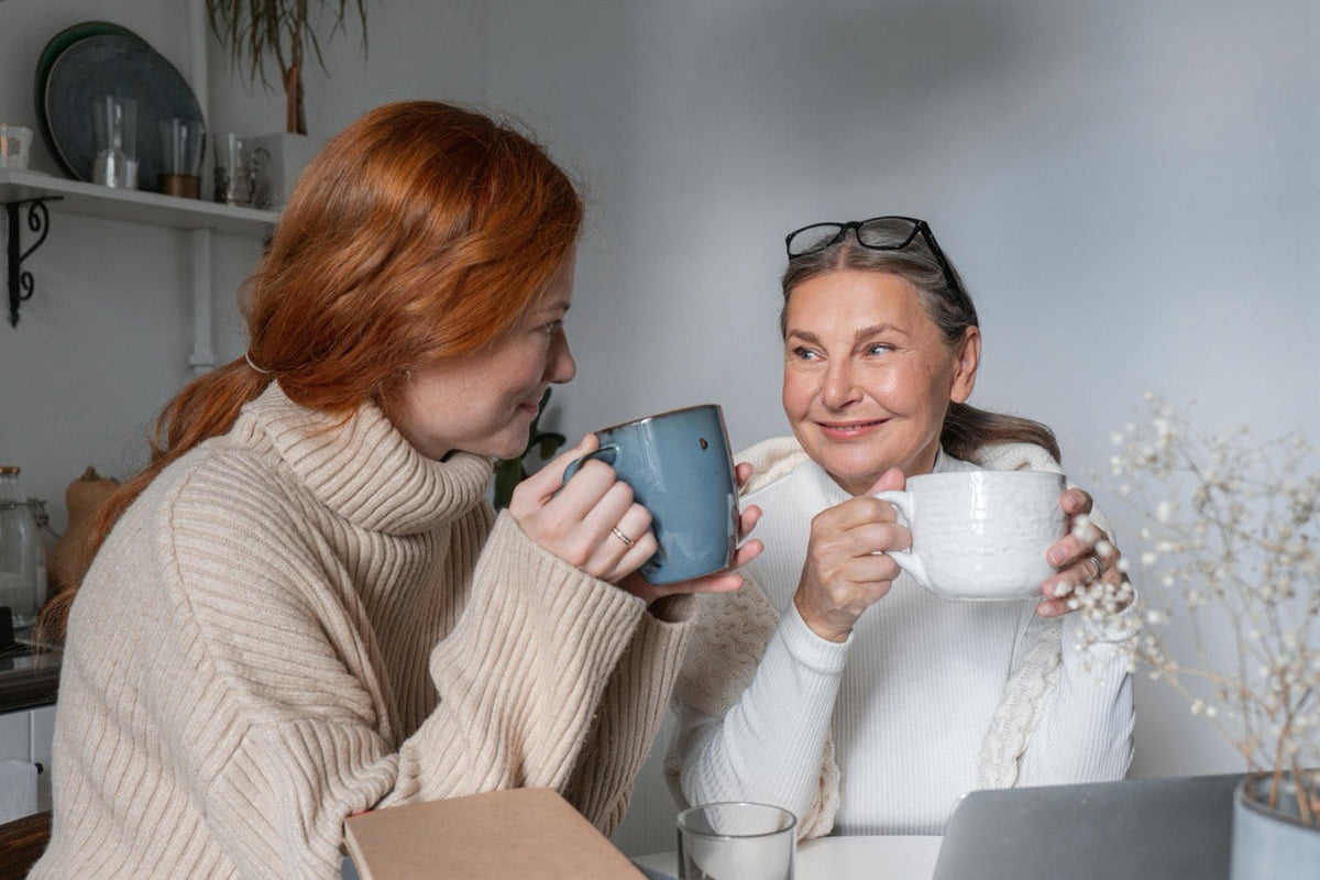 Zwei Frauen sitzen einander zugewandt und schauen sich freundlich an. Beide haben jeweils eine Tasse in der Hand.