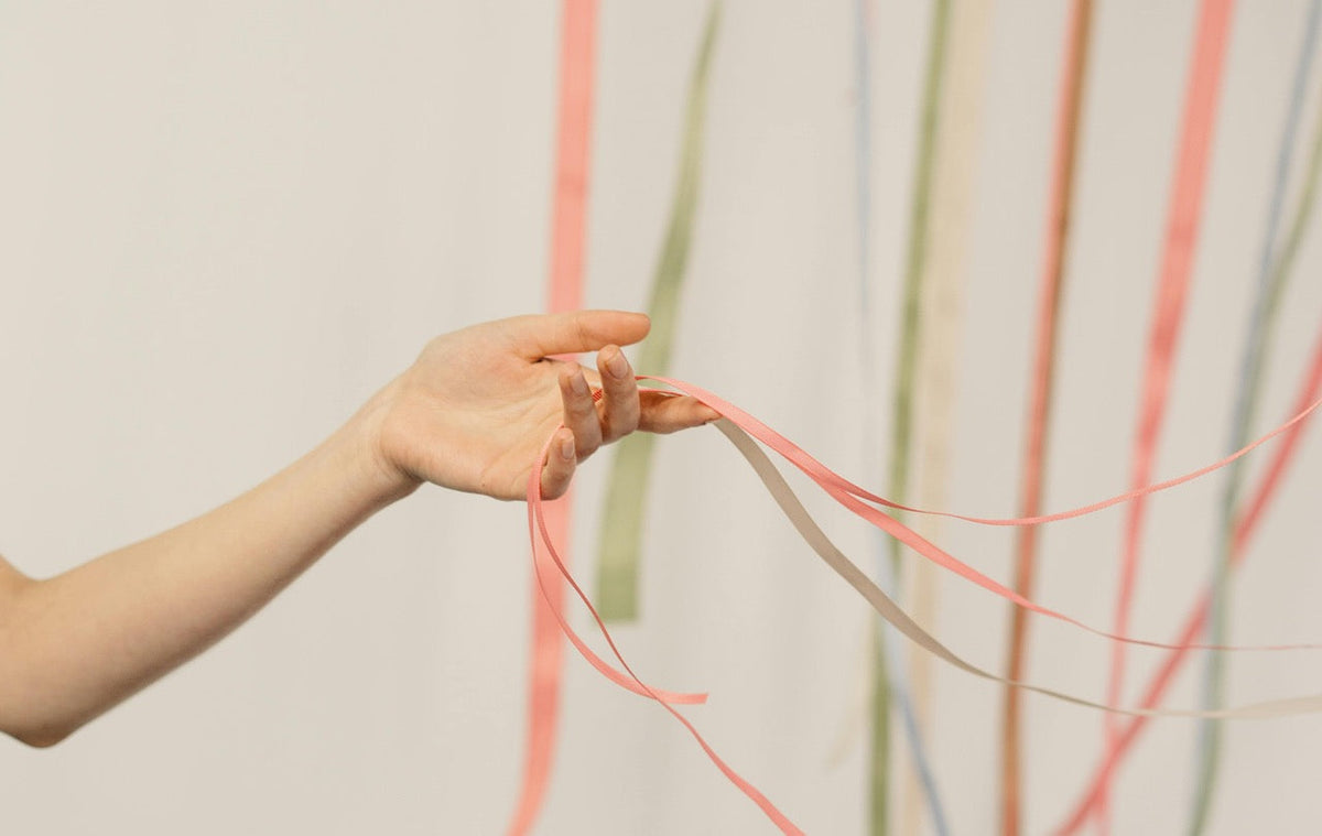 Eine Hand hält zarte, farbige Bänder vor einem hellen Hintergrund. Die Bänder fallen locker herab und vermitteln Leichtigkeit und Bewegung.