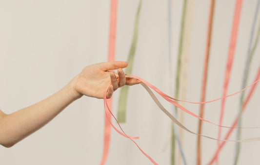 Eine Hand hält zarte, farbige Bänder vor einem hellen Hintergrund. Die Bänder fallen locker herab und vermitteln Leichtigkeit und Bewegung.