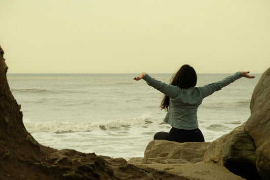 Frau sitzt mit ausgebreiteten Armen am Meer, blickt in die Ferne – ein Moment der Freiheit, Ruhe und inneren Stärke inmitten der Natur.