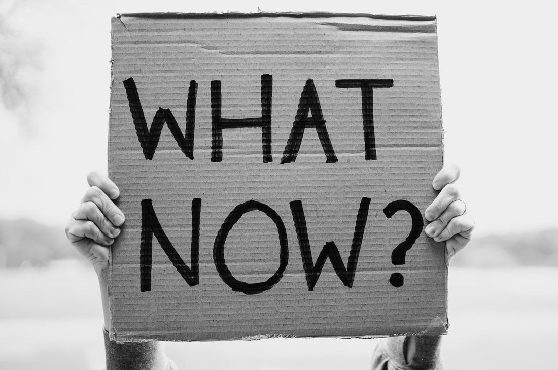 Zwei Hände halten ein Pappschild mit der Aufschrift „WHAT NOW?“ in schwarzer Schrift – als Symbol für Orientierungslosigkeit, Neubeginn oder Entscheidung.