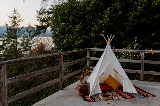 Ein kleines Tipi-Zelt auf einer Terrasse mit Kissen und Decke, umgeben von Wald, Lichterkette und Blick auf Berge und See – ein Ort der Geborgenheit.