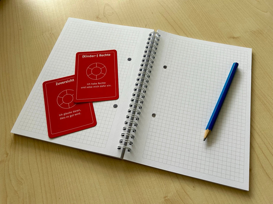 Ein aufgeschlagener Collegeblock liegt auf einem Tisch. Auf der einen Seite liegen zwei orangefarbene Wertekarten, auf der anderen ein blauer Bleistift.