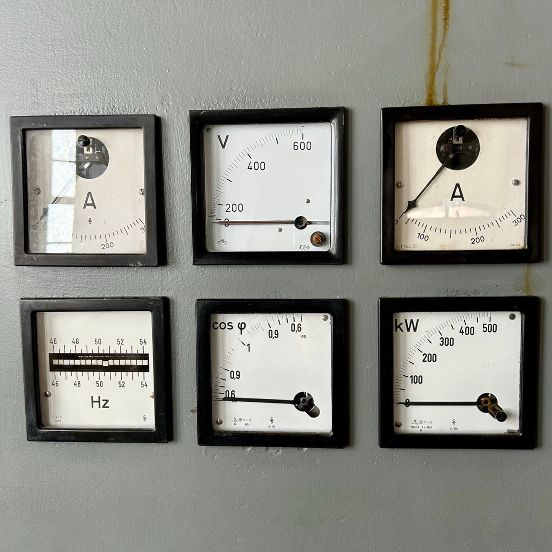 Das Bild zeigt sechs analoge Messinstrumente auf einer grauen Schalttafel. Die Anzeigen messen verschiedene elektrische Parameter.