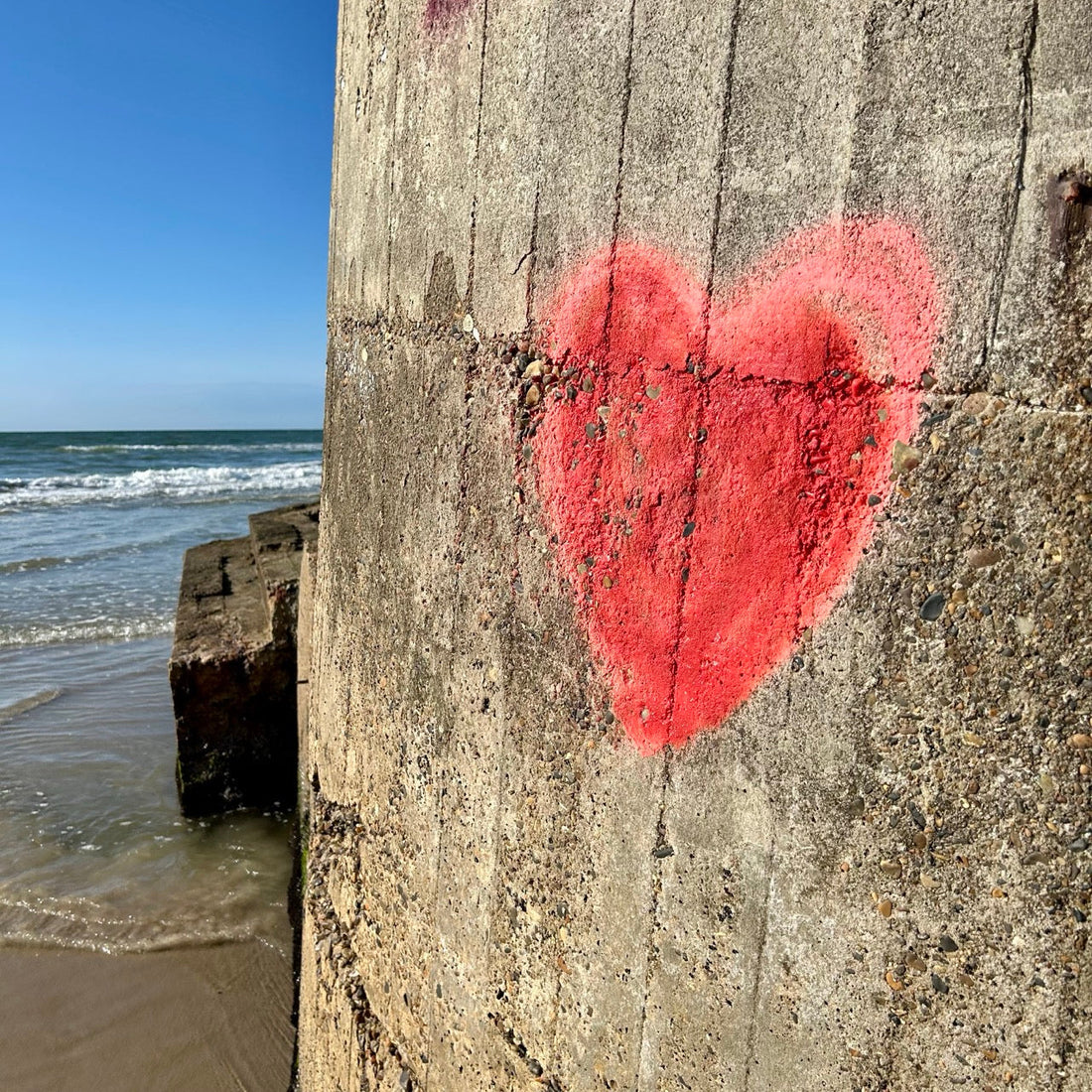 Ein rotes Herz-Graffiti ziert eine raue Betonwand am Strand. Dahinter das Meer mit sanften Wellen unter klarem, blauem Himmel.