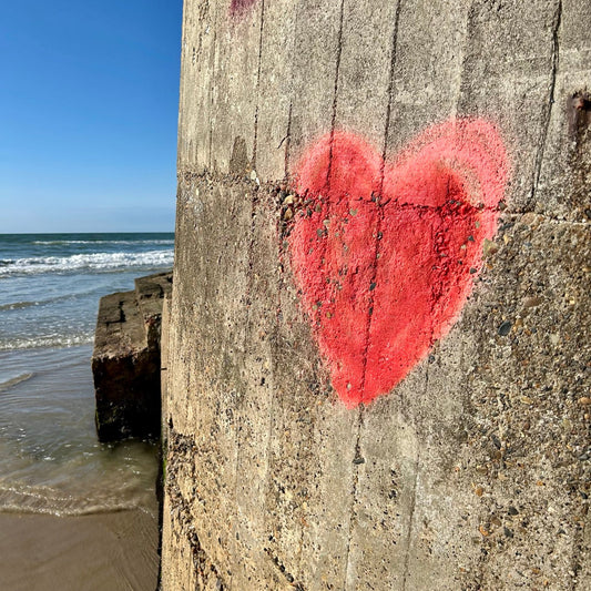 Ein rotes Herz-Graffiti ziert eine raue Betonwand am Strand. Dahinter das Meer mit sanften Wellen unter klarem, blauem Himmel.