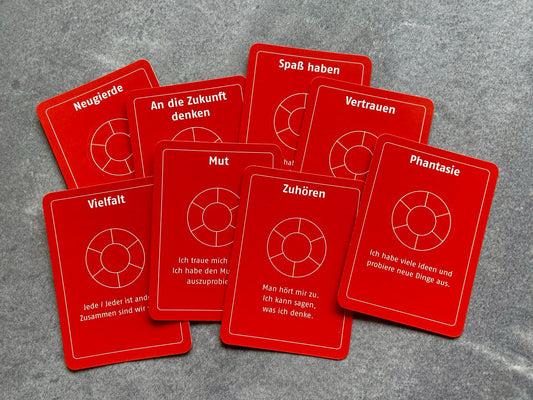 Mehrere rote Wertekarten liegen fächerförmig auf grauem Untergrund. Begriffe wie Vertrauen, Mut, Zuhören und Vielfalt laden zur Reflexion und zum Gespräch ein.
