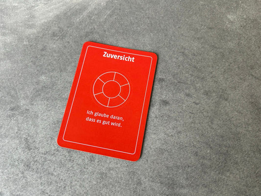 Die orangefarbene Wertekarte "Zuversicht" mit dem Untertitel Ich glaube daran, dass es gut wird" liegt auf einem grauen Boden.