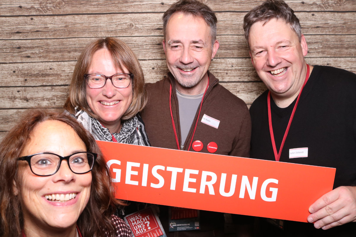 Petra Uhlmann, Imke Meyer, Tim Nieke und Stefan Zollondz sind das Werteteam und halten gemeinsam ein orangefarbenes Schild mit der Aufschrift Begeisterung.